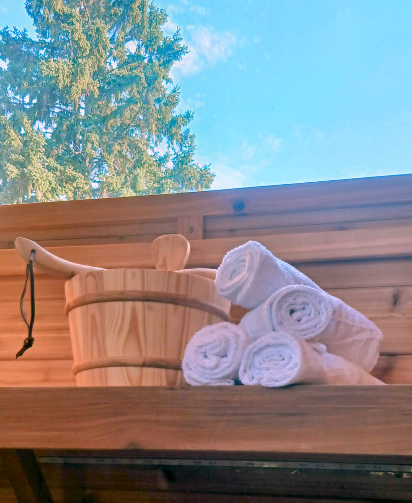 Water pail with wooden ladle and towels waiting to be used on the cedar bench. An evergreen tree and blue sky look picturesque out the window above the bench.